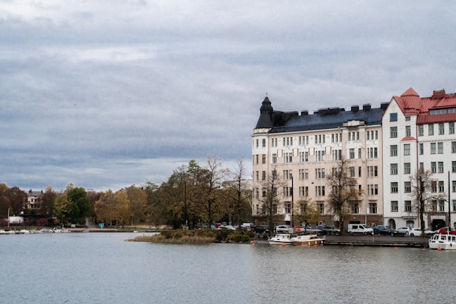 A waterfront scene featuring a row of elegant multi-story buildings with intricate architectural designs. The buildings have varied roof colors including black and red, and sit along a calm body of water. Leafless trees stand in front of the buildings, indicating an autumn setting. Small boats are docked near the shoreline.