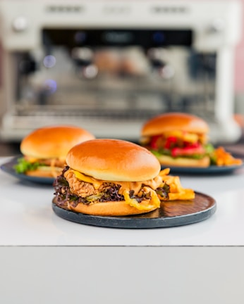 Three gourmet burgers are placed on dark plates on a white surface. The foreground features a burger with a golden bun, crispy lettuce, fried chicken, and a generous portion of creamy sauce overflowing from the side. In the background, two other burgers showcase colorful toppings including red peppers and green lettuce. The blurred backdrop suggests a modern kitchen environment with a blurred appliance.