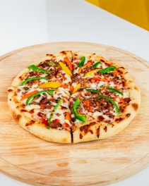 A freshly baked pizza on a wooden serving board, topped with melted cheese, sliced green and yellow bell peppers, diced tomatoes, and ground meat.