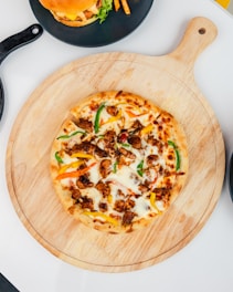 A freshly baked pizza topped with melted cheese, colorful bell peppers, and pieces of grilled meat sits on a wooden board. A cheeseburger with lettuce and fries is on a black plate next to it, all set on a white table.