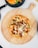 A freshly baked pizza topped with melted cheese, colorful bell peppers, and pieces of grilled meat sits on a wooden board. A cheeseburger with lettuce and fries is on a black plate next to it, all set on a white table.