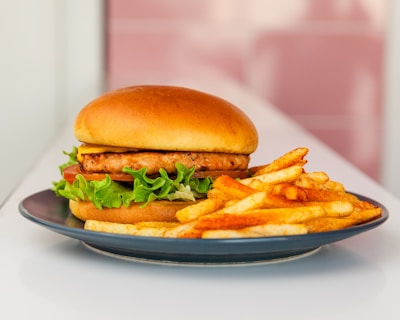 A delicious-looking chicken sandwich sits on a dark plate, accompanied by golden, seasoned fries. The sandwich has a soft, brown bun and is filled with crispy lettuce, a slice of tomato, and a chicken patty.