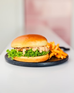 A juicy hamburger placed on a round black plate with a side of golden brown fries. The burger is layered with fresh green lettuce and a generous amount of creamy sauce, all encased within a soft, lightly toasted bun.