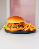 A delicious-looking chicken burger with crispy fried chicken, fresh lettuce, and sauce is placed on a round black plate. Next to the burger are golden-brown seasoned fries. The background is softly blurred, drawing attention to the food.