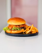 A delicious-looking chicken burger with crispy fried chicken, fresh lettuce, and sauce is placed on a round black plate. Next to the burger are golden-brown seasoned fries. The background is softly blurred, drawing attention to the food.