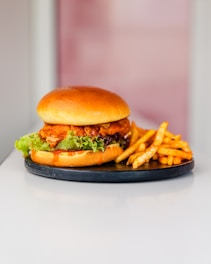 A delicious-looking chicken burger with crispy fried chicken, fresh lettuce, and sauce is placed on a round black plate. Next to the burger are golden-brown seasoned fries. The background is softly blurred, drawing attention to the food.