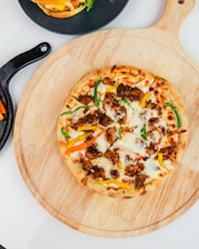 A freshly baked pizza with a variety of toppings including melted cheese, green and yellow bell peppers, and pieces of meat is placed on a wooden serving board. In the background, there is a partially visible hamburger on a black plate and a dish containing fries or another side item.