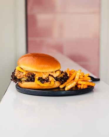 A gourmet burger with a glossy bun is served on a black plate. It features crispy fried chicken, lettuce, and a generous amount of sauce. Beside the burger, there is a serving of golden, seasoned fries. The background includes a soft, pink tiled wall.