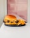 A gourmet burger with a glossy bun is served on a black plate. It features crispy fried chicken, lettuce, and a generous amount of sauce. Beside the burger, there is a serving of golden, seasoned fries. The background includes a soft, pink tiled wall.