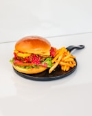 A freshly prepared burger with a glossy, golden-brown bun sits on a round, black serving plate. The burger is filled with layers of vibrant green lettuce, juicy tomato slices, crispy fried chicken, melted cheese, and bright red peppers. A generous portion of golden-brown seasoned fries is placed beside the burger, complementing the meal.