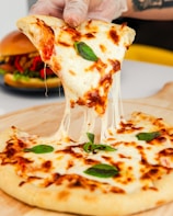 A cheesy slice of margherita pizza being lifted from the wooden serving board, showcasing stretchy mozzarella cheese with fresh basil leaves on top. In the background, there is a blurred image of a burger with visible red peppers and lettuce.
