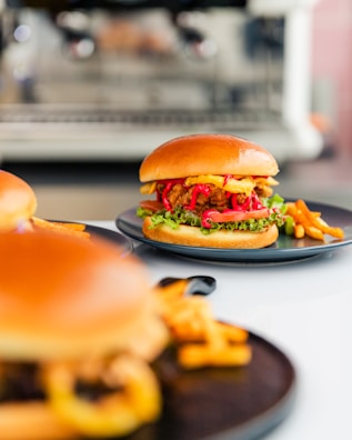 A close-up view of a delicious-looking burger with a glossy bun, crispy fried chicken, fresh lettuce, vibrant red onions, and a drizzle of sauce, served on a dark plate alongside golden fries. The background is softly blurred, highlighting the appetizing meal.