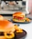 A close-up view of a delicious-looking burger with a glossy bun, crispy fried chicken, fresh lettuce, vibrant red onions, and a drizzle of sauce, served on a dark plate alongside golden fries. The background is softly blurred, highlighting the appetizing meal.
