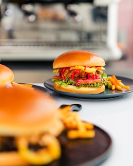 A close-up view of a delicious-looking burger with a glossy bun, crispy fried chicken, fresh lettuce, vibrant red onions, and a drizzle of sauce, served on a dark plate alongside golden fries. The background is softly blurred, highlighting the appetizing meal.