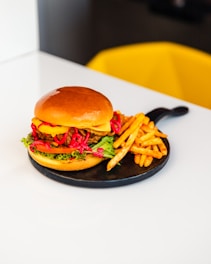 A juicy cheeseburger topped with sliced tomatoes, lettuce, cheddar cheese, red onions, and vibrant red peppers sits on a black platter alongside a serving of crispy golden fries. The setting is a bright white tabletop, with a hint of a vibrant yellow chair in the background.