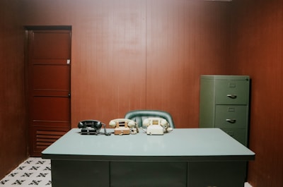 A minimalist office setting with a gray desk and a vintage telephone setup. The room features a dark wood-paneled wall, a green filing cabinet, and a simple chair. The design reflects a retro or mid-century aesthetic.
