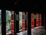 A traditional architecture scene featuring open wooden doors with ornate designs. The building features rich, dark colors with prominent red and gold accents on the doors. Outside, a lush garden with various plants, including a palm tree, can be seen through the doorways. The tiled floor enhances the older, historical feel of the location.