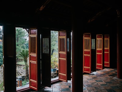 A traditional architecture scene featuring open wooden doors with ornate designs. The building features rich, dark colors with prominent red and gold accents on the doors. Outside, a lush garden with various plants, including a palm tree, can be seen through the doorways. The tiled floor enhances the older, historical feel of the location.