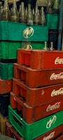 Stacks of green and red plastic crates, with empty glass bottles. The crates are branded with Pepsi and Coca-Cola logos. The bottles are clear and placed in a disorganized manner.