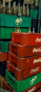 Stacks of green and red plastic crates, with empty glass bottles. The crates are branded with Pepsi and Coca-Cola logos. The bottles are clear and placed in a disorganized manner.