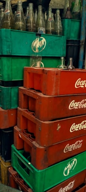 Stacks of green and red plastic crates, with empty glass bottles. The crates are branded with Pepsi and Coca-Cola logos. The bottles are clear and placed in a disorganized manner.