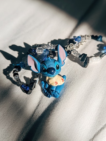 A small, blue cartoon character figurine with large ears and a mischievous expression is placed on a soft, light-colored surface. Surrounding the figurine are beaded bracelets incorporating blue, white, and black beads, with some clear crystal-like beads as well. The lighting creates distinct shadows and highlights on both the figurine and the bracelets.
