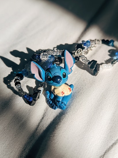 A small, blue cartoon character figurine with large ears and a mischievous expression is placed on a soft, light-colored surface. Surrounding the figurine are beaded bracelets incorporating blue, white, and black beads, with some clear crystal-like beads as well. The lighting creates distinct shadows and highlights on both the figurine and the bracelets.