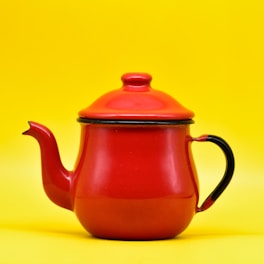 A vibrant red teapot with a glossy finish is positioned against a bright yellow background. The teapot features a curved spout, a handle, and a lid with a small knob on top.