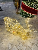A decorative reindeer sculpture is adorned with glowing fairy lights, positioned on a cobblestone pathway. Nearby, a large, illuminated cone shape with red and green lights suggests a festive holiday theme. Two people stand in the background, partially visible.