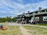 A rustic, two-story wooden building with slightly worn brown siding and green-trimmed windows sits as the central focus. Several people with backpacks and outdoor gear gather near the entrance, suggesting an atmosphere of camaraderie and adventure. In the foreground, a path leads to the building, bordered by grass and a wooden bench. The sky is overcast with patches of blue, and the surrounding landscape features sparse trees, adding a sense of openness.