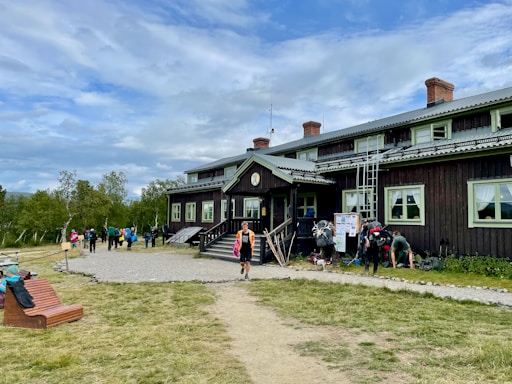 A rustic, two-story wooden building with slightly worn brown siding and green-trimmed windows sits as the central focus. Several people with backpacks and outdoor gear gather near the entrance, suggesting an atmosphere of camaraderie and adventure. In the foreground, a path leads to the building, bordered by grass and a wooden bench. The sky is overcast with patches of blue, and the surrounding landscape features sparse trees, adding a sense of openness.