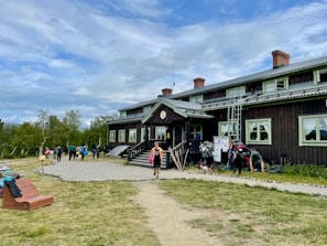 A rustic, two-story wooden building with slightly worn brown siding and green-trimmed windows sits as the central focus. Several people with backpacks and outdoor gear gather near the entrance, suggesting an atmosphere of camaraderie and adventure. In the foreground, a path leads to the building, bordered by grass and a wooden bench. The sky is overcast with patches of blue, and the surrounding landscape features sparse trees, adding a sense of openness.