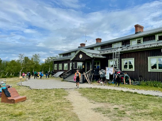 A rustic, two-story wooden building with slightly worn brown siding and green-trimmed windows sits as the central focus. Several people with backpacks and outdoor gear gather near the entrance, suggesting an atmosphere of camaraderie and adventure. In the foreground, a path leads to the building, bordered by grass and a wooden bench. The sky is overcast with patches of blue, and the surrounding landscape features sparse trees, adding a sense of openness.