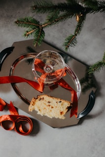A metallic serving tray holds a slice of a fruit-studded cake and a glass with a red ribbon tied around the stem. Sprigs of evergreen branches extend over the tray, adding a festive touch. Red ribbons are loosely arranged around the tray, contributing to a holiday-themed setup.