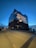 A large, modern concert hall features a unique wave-like glass structure on top of a brick base. The building is illuminated, creating a striking contrast against the darkening blue sky. People walk along a path leading up to the building, and there are city and water elements in the background.
