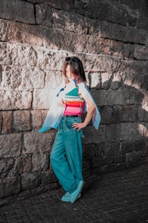 A person stands against a stone brick wall, wearing a colorful striped tank top, loose blue jeans, and light-colored shoes. sunlight creates a shadow pattern on the wall, highlighting the person's hair and partially covering their face. The relaxed pose and casual attire suggest a casual or leisurely mood.