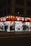A well-lit street-side shop at night featuring bright orange signage and a variety of merchandise displayed outside, including clothing, accessories, and household items. Several people walk past the store along the sidewalk, casting shadows on the pavement.