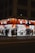 A well-lit street-side shop at night featuring bright orange signage and a variety of merchandise displayed outside, including clothing, accessories, and household items. Several people walk past the store along the sidewalk, casting shadows on the pavement.