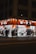 A well-lit street-side shop at night featuring bright orange signage and a variety of merchandise displayed outside, including clothing, accessories, and household items. Several people walk past the store along the sidewalk, casting shadows on the pavement.