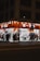 A well-lit street-side shop at night featuring bright orange signage and a variety of merchandise displayed outside, including clothing, accessories, and household items. Several people walk past the store along the sidewalk, casting shadows on the pavement.