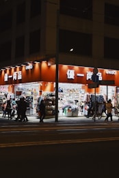 A well-lit street-side shop at night featuring bright orange signage and a variety of merchandise displayed outside, including clothing, accessories, and household items. Several people walk past the store along the sidewalk, casting shadows on the pavement.