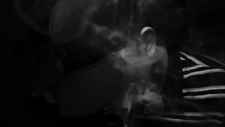 A person wearing a baseball cap is partially obscured by swirling smoke. The person is in a dark setting, with minimal light highlighting parts of their ear and a striped shirt they are wearing.