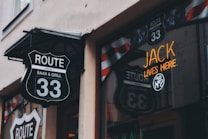 The image features the exterior of an establishment named 'Route 33 Baar & Grill.' There is a distinctive black and white route shield sign hanging above the entrance door. Additionally, a reflection of the same sign is visible in the window. The window also displays a neon sign with the words 'Jack lives here.' In the background, there are patriotic banners with stars and stripes.