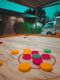 A carrom board with colorful striker and coins is placed on a table. The scene is set outdoors under a canopy with a view of a lush garden and a white van in the background.