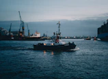 A powerful tugboat assisting a large cargo ship into the busy London Medway port under a cloudy sky.