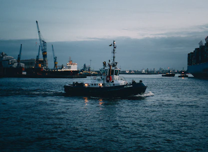 A sturdy tug boat maneuvering a large vessel near a busy port under clear skies.