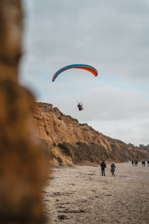 A paraglider with a blue and orange canopy is soaring above a sandy beach with cliffs in the background. Below, a few people are walking along the shoreline under a cloudy sky.