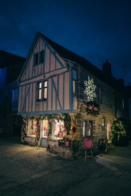 A quaint, timber-framed building adorned with festive decorations under a dark evening sky. It has illuminated windows showcasing warm, inviting displays, with Christmas lights and wreaths adding to the holiday ambiance. A lit snowflake decoration is projected on the wall.