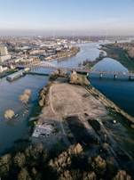 Aerial view of a river landscape with both urban and natural elements. A long bridge spans across the river, connecting two sides of a city. On one side is a densely built urban area with buildings and industrial structures, while the other side is dominated by open green space and patches of bare land. Trees, some of which appear to be flooded, are visible along the riverbanks. Wind turbines can be seen in the distance under a clear blue sky.