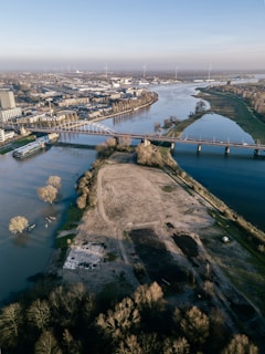 Aerial view of a river landscape with both urban and natural elements. A long bridge spans across the river, connecting two sides of a city. On one side is a densely built urban area with buildings and industrial structures, while the other side is dominated by open green space and patches of bare land. Trees, some of which appear to be flooded, are visible along the riverbanks. Wind turbines can be seen in the distance under a clear blue sky.